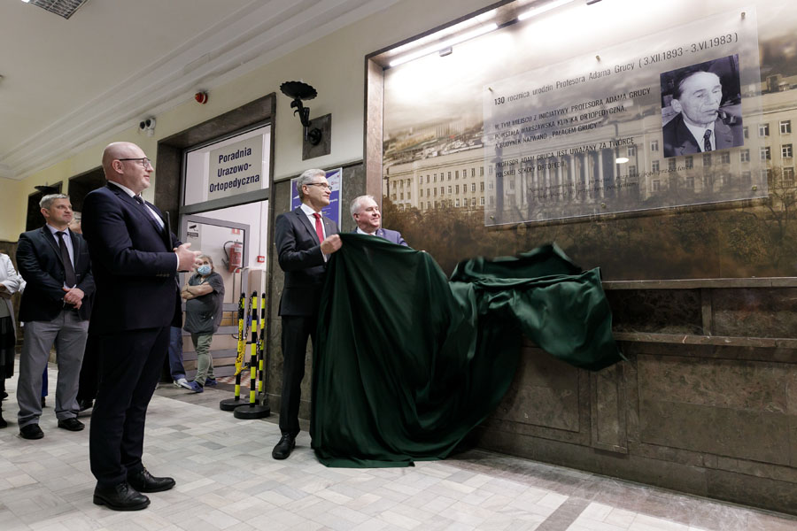 Na zdjęciu kilka osób, stoją przed tablicą powieszoną na ścianie. Dwóch mężczyzn stojących pod tablicą trzyma zielone sukno.
